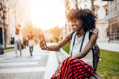 Genç siyah bayan turist güzel bir Avrupa şehrinin sokaklarında yürümekten zevk alıyor. O mutlu ve akıllı telefonunu bazı fotoğraflar ve videolar çekmek için kullanıyor. Parlak güneşli bir gün.