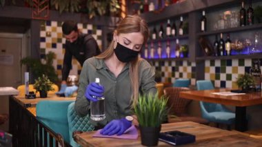 Young restaurant waiters cleaning and disinfecting tables and surfaces during Coronavirus pandemic disease. They are wearing protective face masks and gloves.
