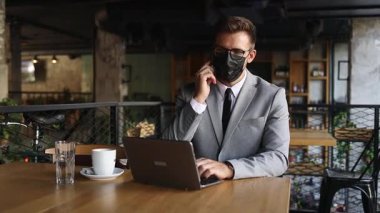 Yüzünde koruyucu maske olan bir iş adamı bir kafede oturuyor ve dizüstü bilgisayar kullanırken telefonla konuşuyor. Coronavirus, Covid-19 konsepti.
