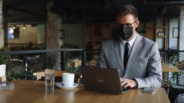 Yüzünde koruyucu maske olan bir iş adamı bir kafede oturuyor ve dizüstü bilgisayar kullanırken telefonla konuşuyor. Coronavirus, Covid-19 konsepti.