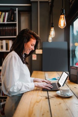 Güzel bir bayan öğrenci ya da genç bir iş kadını dizüstü bilgisayarını kullanıyor ve rahat bir kafede pencerenin yanında duruyor..