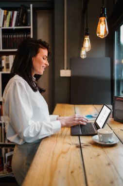 Güzel bir bayan öğrenci ya da genç bir iş kadını dizüstü bilgisayarını kullanıyor ve rahat bir kafede pencerenin yanında duruyor..