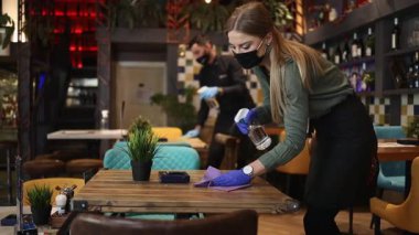 Genç restoran garsonları Coronavirus salgını sırasında masaları ve yüzeyleri temizleyip dezenfekte ediyorlar. Koruyucu maske ve eldiven takıyorlar..