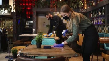 Genç restoran garsonları Coronavirus salgını sırasında masaları ve yüzeyleri temizleyip dezenfekte ediyorlar. Koruyucu maske ve eldiven takıyorlar..