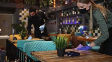 Genç restoran garsonları Coronavirus salgını sırasında masaları ve yüzeyleri temizleyip dezenfekte ediyorlar. Koruyucu maske ve eldiven takıyorlar..