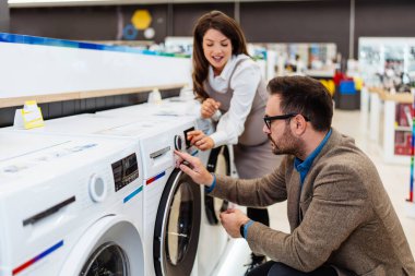 Beautiful and happy middle age couple buying consumer tech products in modern home appliances store. They are choosing washing or cloth dryer machine. People and consumerism concept.