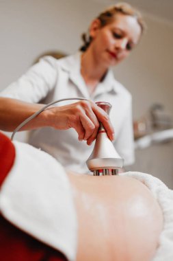 Güzellik uzmanı, profesyonel güzellik merkezinde yatan gülümseyen olgun bir kadının karnına radyasyon kaldırma cihazı uyguluyor. Modern, zararsız bir vücut şekillendirme tedavisi sunuyor..