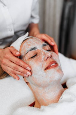 Profesyonel güzellik uzmanı gevşemiş bir müşterinin yüzüne nazikçe kremsi bir maske takarak sağlıklı cilt için rahatlatıcı ve canlandırıcı bir spa tedavisi uyguluyor.