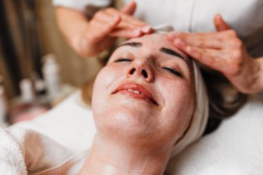 Profesyonel güzellik uzmanı gevşemiş bir müşterinin yüzüne nazikçe kremsi bir maske takarak sağlıklı cilt için rahatlatıcı ve canlandırıcı bir spa tedavisi uyguluyor.