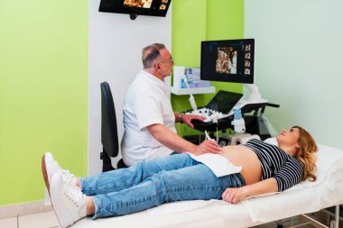 Modern ultrason tarayıcısı kullanan jinekolog, tıbbi muayene masasında yatan hamile bir kadın üzerinde 4D tarama yapıyor.