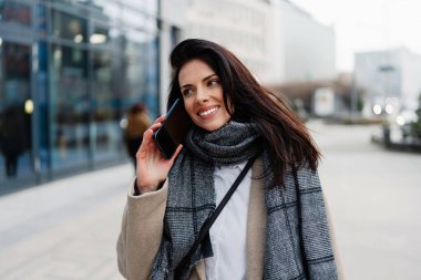 Şık bir ceket ve eşarpla gülümseyen iş kadını modern bir ofis binasının önünde akıllı telefonuyla selfie çekiyor, kentsel bağlantıların ve kişisel markaların tadını çıkarıyor.