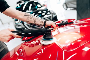 Profesyonel araba temizleyicisi kırmızı bir arabanın kaputunu cilalamak için yörünge cilası kullanıyor. Modern bir oto tamirhanesinde pürüzsüz ve parlak bir son sağlıyor.