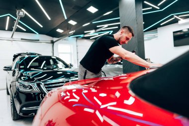 Profesyonel araba temizleyicisi kırmızı bir arabanın kaputunu cilalamak için yörünge cilası kullanıyor. Modern bir oto tamirhanesinde pürüzsüz ve parlak bir son sağlıyor.