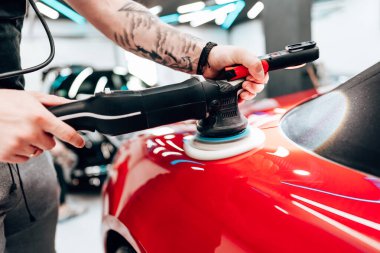 Profesyonel araba temizleyicisi kırmızı bir arabanın kaputunu cilalamak için yörünge cilası kullanıyor. Modern bir oto tamirhanesinde pürüzsüz ve parlak bir son sağlıyor.