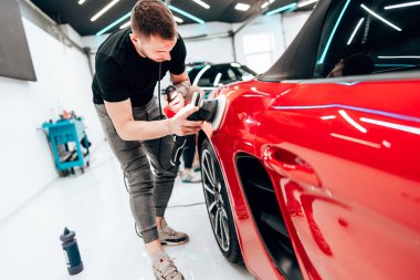 Profesyonel araba temizleyicisi kırmızı bir arabanın kaputunu cilalamak için yörünge cilası kullanıyor. Modern bir oto tamirhanesinde pürüzsüz ve parlak bir son sağlıyor.