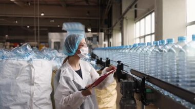 Koruyucu giysiler ve maske takan kadın kalite kontrol müfettişi su şişeleme fabrikasındaki üretim hattını incelerken panoya notlar yazıyor.