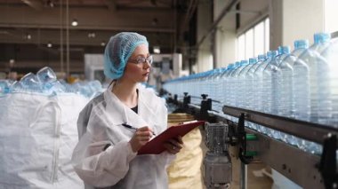 Koruyucu giysiler ve maske takan kadın kalite kontrol müfettişi su şişeleme fabrikasındaki üretim hattını incelerken panoya notlar yazıyor.
