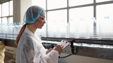 Koruyucu giysiler ve maske takan kadın kalite kontrol müfettişi su şişeleme fabrikasındaki üretim hattını incelerken panoya notlar yazıyor.