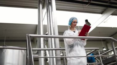 Koruyucu iş kıyafetlerinde çalışan, tıbbi malzeme araştırma ve üretim fabrikasında çalışan kadın işçi. Kontrol köprüsünde duruyor veya yürüyor ve üretim hattını izliyor..