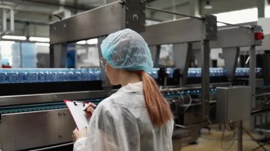Koruyucu giysiler ve maske takan kadın kalite kontrol müfettişi su şişeleme fabrikasındaki üretim hattını incelerken panoya notlar yazıyor.