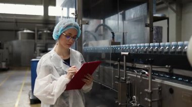 Koruyucu giysiler ve maske takan kadın kalite kontrol müfettişi su şişeleme fabrikasındaki üretim hattını incelerken panoya notlar yazıyor.