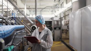 Koruyucu giysiler ve maske takan kadın kalite kontrol müfettişi su şişeleme fabrikasındaki üretim hattını incelerken panoya notlar yazıyor.