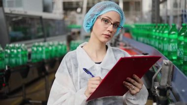 Koruyucu giysiler ve maske takan kadın kalite kontrol müfettişi su şişeleme fabrikasındaki üretim hattını incelerken panoya notlar yazıyor.