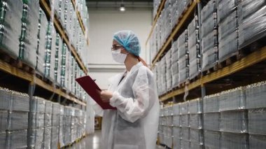 Dağıtım deposundaki raflarda depolanan ürün ve eşyaları incelerken, laboratuvar önlüğü ve saç filesi ile panoya notlar yazan kadın denetçi.