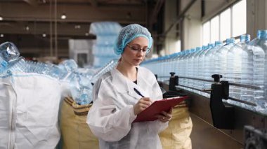 Koruyucu giysiler ve maske takan kadın kalite kontrol müfettişi su şişeleme fabrikasındaki üretim hattını incelerken panoya notlar yazıyor.
