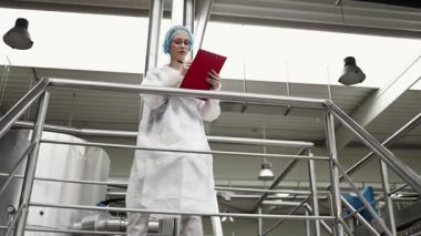 Koruyucu iş kıyafetlerinde çalışan, tıbbi malzeme araştırma ve üretim fabrikasında çalışan kadın işçi. Kontrol köprüsünde duruyor veya yürüyor ve üretim hattını izliyor..