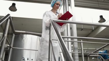 Koruyucu iş kıyafetlerinde çalışan, tıbbi malzeme araştırma ve üretim fabrikasında çalışan kadın işçi. Kontrol köprüsünde duruyor veya yürüyor ve üretim hattını izliyor..