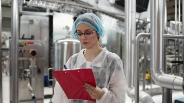 Koruyucu giysiler ve maske takan kadın kalite kontrol müfettişi su şişeleme fabrikasındaki üretim hattını incelerken panoya notlar yazıyor.