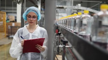 Koruyucu giysiler ve maske takan kadın kalite kontrol müfettişi su şişeleme fabrikasındaki üretim hattını incelerken panoya notlar yazıyor.