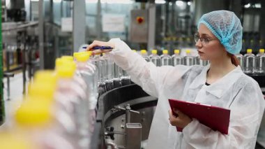 Koruyucu giysiler ve maske takan kadın kalite kontrol müfettişi su şişeleme fabrikasındaki üretim hattını incelerken panoya notlar yazıyor.