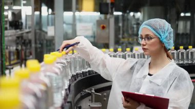 Koruyucu giysiler ve maske takan kadın kalite kontrol müfettişi su şişeleme fabrikasındaki üretim hattını incelerken panoya notlar yazıyor.