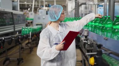 Koruyucu giysiler ve maske takan kadın kalite kontrol müfettişi su şişeleme fabrikasındaki üretim hattını incelerken panoya notlar yazıyor.