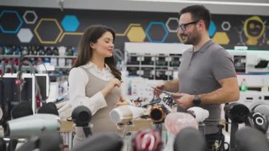 Neşeli bir elektronik mağazasında çeşitli ürünler sergilerken hamile bir müşteriye ev aletleri seçmesinde yardımcı olan gülümseyen mağaza asistanı