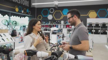 Neşeli bir elektronik mağazasında çeşitli ürünler sergilerken hamile bir müşteriye ev aletleri seçmesinde yardımcı olan gülümseyen mağaza asistanı