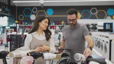 Neşeli bir elektronik mağazasında çeşitli ürünler sergilerken hamile bir müşteriye ev aletleri seçmesinde yardımcı olan gülümseyen mağaza asistanı