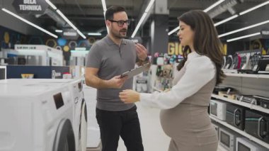 Elinde tablet olan gülümseyen bir satıcı hamile bir müşteriye çamaşır makinesini gösteriyor ve tartışıyor. Modern bir ev aletleri mağazasından en iyi seçenekleri seçiyor.