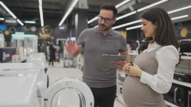 Elinde tablet olan gülümseyen bir satıcı hamile bir müşteriye çamaşır makinesini gösteriyor ve tartışıyor. Modern bir ev aletleri mağazasından en iyi seçenekleri seçiyor.