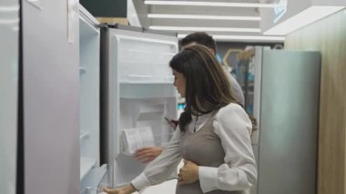 Elinde tabletle gülümseyen bir satıcı hamile bir müşteriye buzdolabını gösteriyor ve tartışıyor. Modern bir ev aletleri mağazasından en iyi seçenekleri seçiyor.