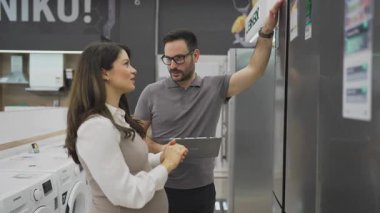 Elinde tabletle gülümseyen bir satıcı hamile bir müşteriye buzdolabını gösteriyor ve tartışıyor. Modern bir ev aletleri mağazasından en iyi seçenekleri seçiyor.