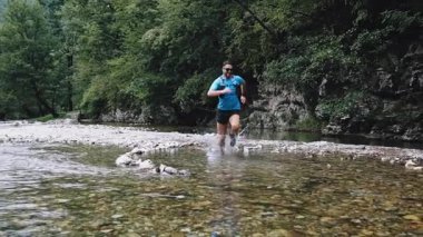 Zengin bir ormanda nehirde koşan erkek sporcu, su sıçratıyor ve doğanın güzelliği arasında bir patika boyunca azim ve atletizm sergiliyor.