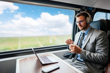 Kulaklık ve takım elbiseli genç işadamı, tren yolculuğu boyunca bir fincan kahve içerken dizüstü bilgisayarında bir video konferansı yapıyor.