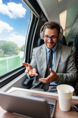 Kulaklık ve takım elbiseli genç işadamı, tren yolculuğu boyunca bir fincan kahve içerken dizüstü bilgisayarında bir video konferansı yapıyor.
