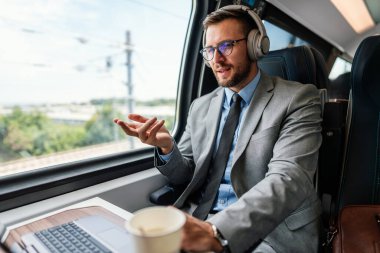 Kulaklık ve takım elbiseli genç işadamı, tren yolculuğu boyunca bir fincan kahve içerken dizüstü bilgisayarında bir video konferansı yapıyor.