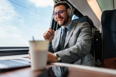 Kulaklık ve takım elbiseli genç işadamı, tren yolculuğu boyunca bir fincan kahve içerken dizüstü bilgisayarında bir video konferansı yapıyor.