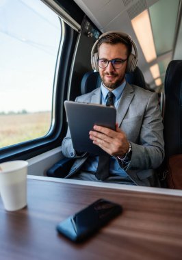 Genç işadamı kulaklık takıyor ve trenle seyahat ederken dijital tablet kullanıyor. Müzikten zevk alıyor ve iş seyahati sırasında üretken kalıyor.