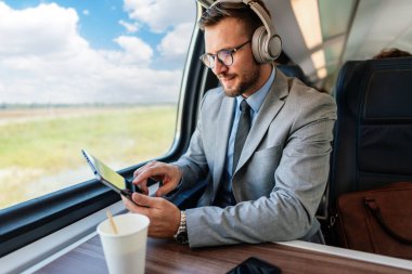 Genç işadamı kulaklık takıyor ve trenle seyahat ederken dijital tablet kullanıyor. Müzikten zevk alıyor ve iş seyahati sırasında üretken kalıyor.